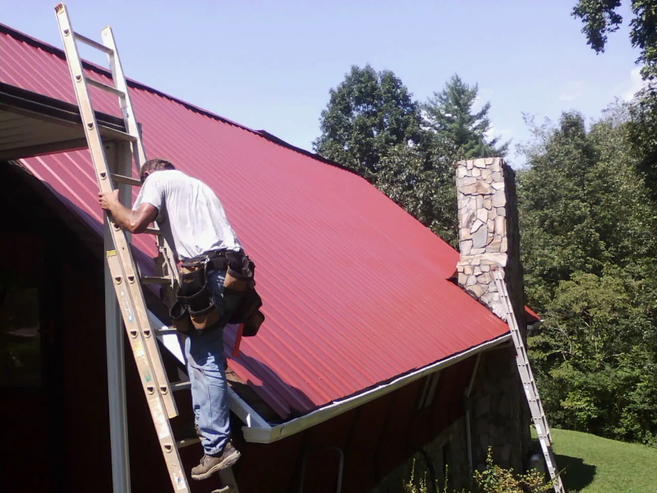 Roofer on a ladder beside a red metal roof and stone chimney — Soffit & Fascia Repair by Roofing Brothers in Apison